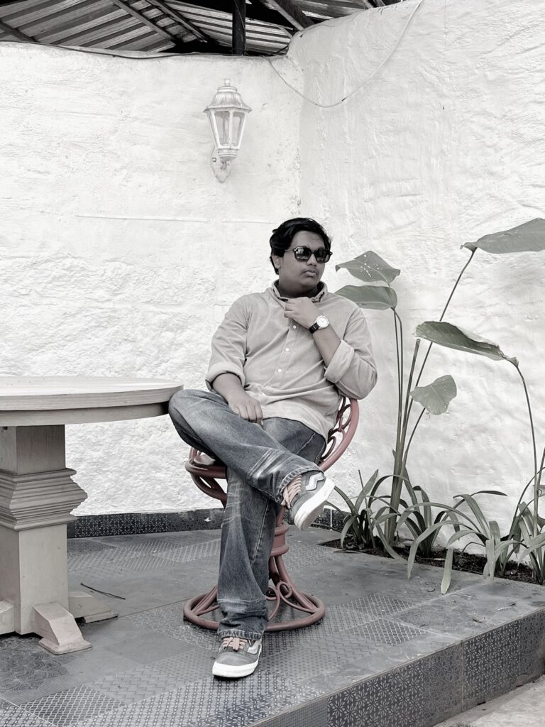 A man siting outdoors at night near a rustic building wall with large windows. He is wearing a checkered shirt with visible patches and has one hand in his pocket. The scene is dimly lit, with plants and wooden benches on the left side and a signboard above him. The photo is in black and white, creating a dramatic, moody atmosphere.