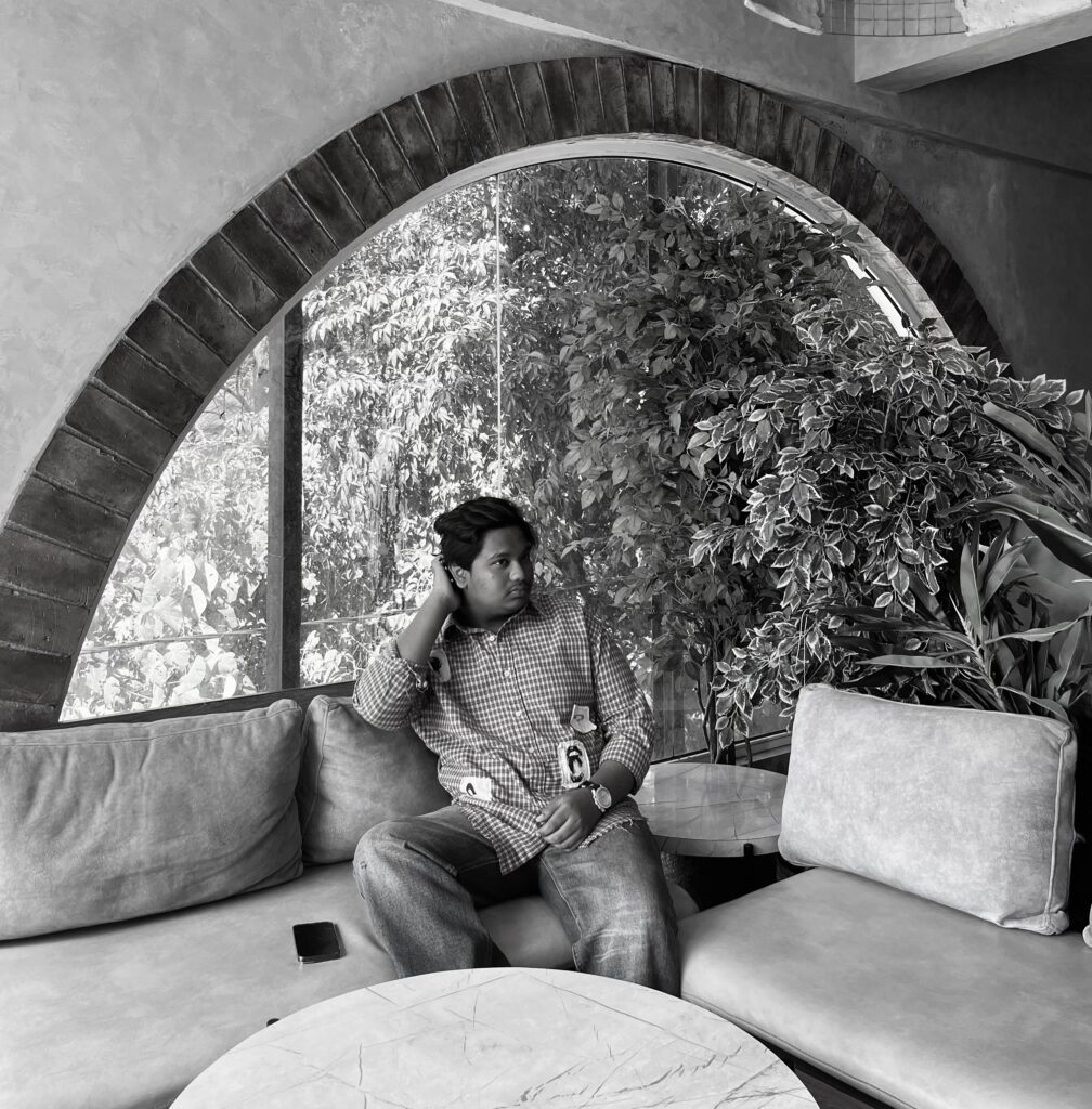 A man stands siting outdoors at night near a rustic building wall with large windows. He is wearing a checkered shirt with visible patches and has one hand in his pocket. The scene is dimly lit, with plants and wooden benches on the left side and a signboard above him. The photo is in black and white, creating a dramatic, moody atmosphere.