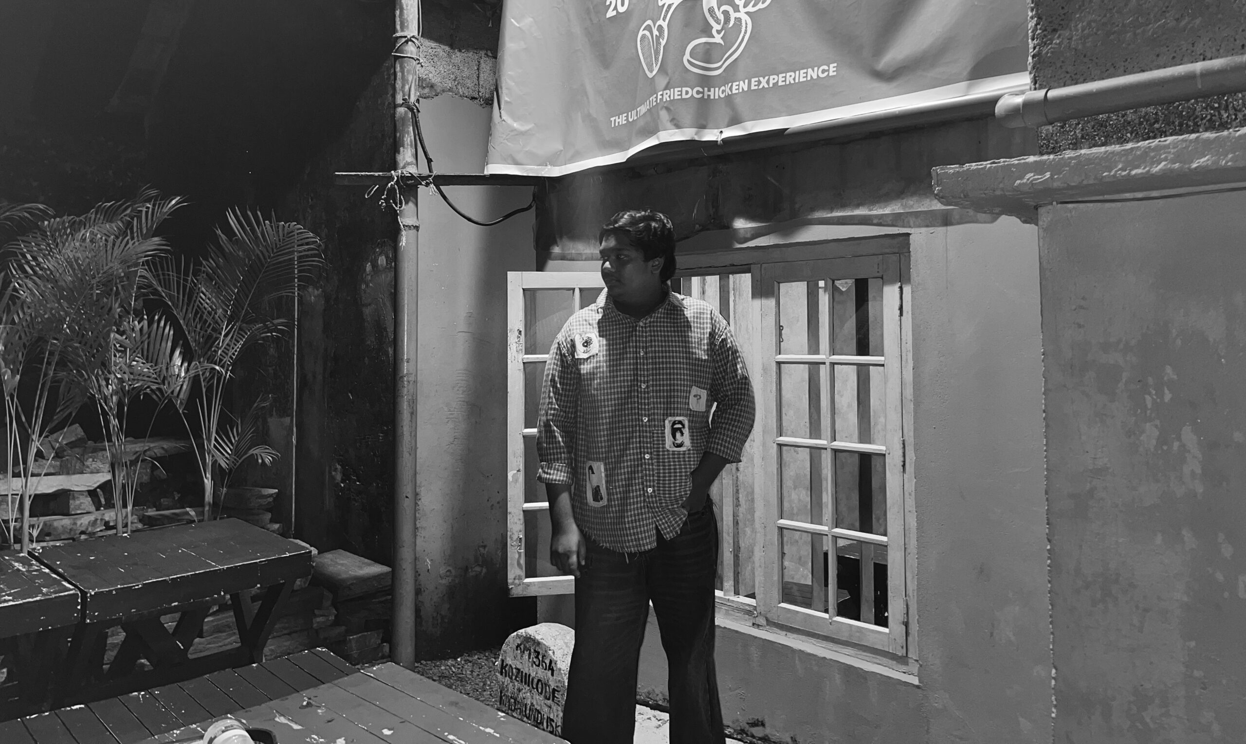 A man stands outdoors at night near a rustic building wall with large windows. He is wearing a checkered shirt with visible patches and has one hand in his pocket. The scene is dimly lit, with plants and wooden benches on the left side and a signboard above him. The photo is in black and white, creating a dramatic, moody atmosphere.