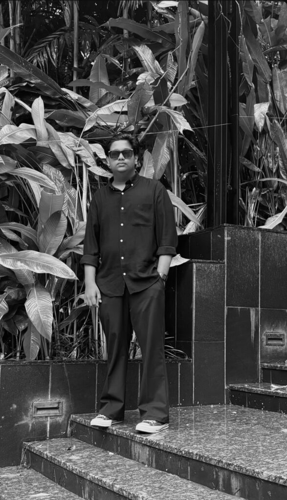 A man stands outdoors at night near a rustic building wall with large windows. He is wearing a checkered shirt with visible patches and has one hand in his pocket. The scene is dimly lit, with plants and wooden benches on the left side and a signboard above him. The photo is in black and white, creating a dramatic, moody atmosphere.
